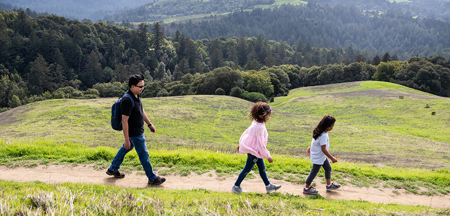 A parent and two kids walk on a dirt trail surrounded by green hillsides.